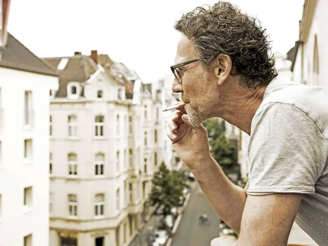 Mann mit Brille raucht auf Balkon, während Zigarettenrauch in Wohnung zieht. Im Hintergrund Häuserfassaden und Straße.