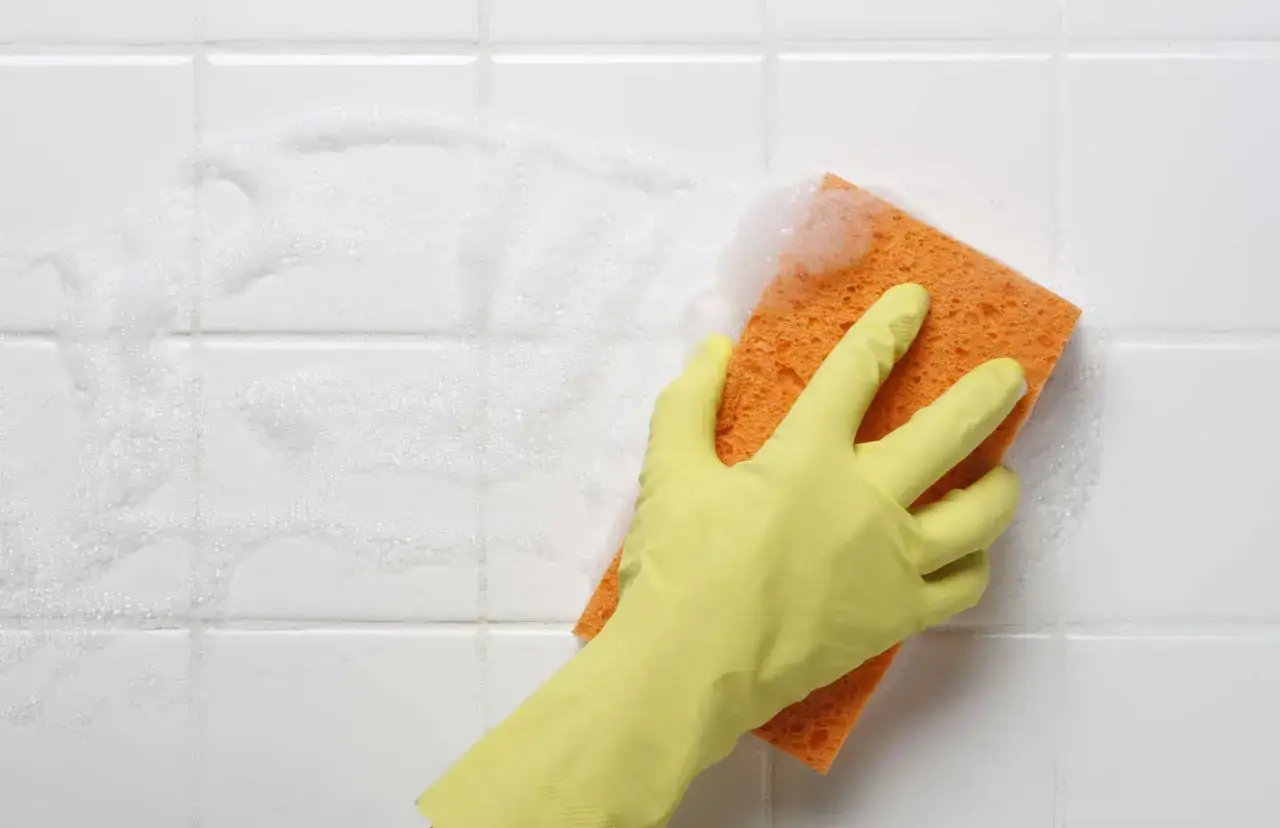 Gelber Handschuh mit orangefarbenem Schwamm reinigt schaumige weiße Wandfliesen.