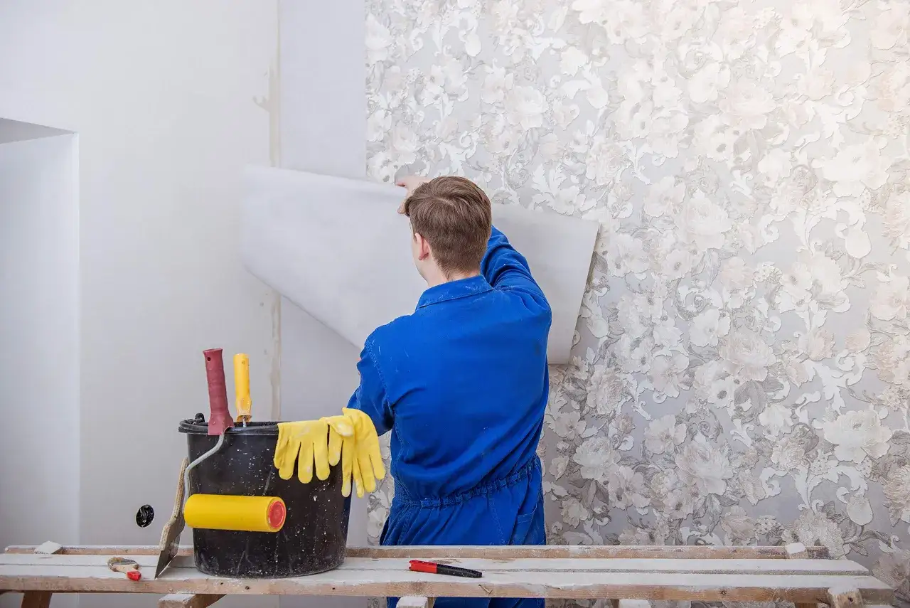 Mann in blauer Latzhose tapeziert eine Wand mit Blumenmuster. Werkzeuge wie Rollen und Handschuhe liegen bereit. Die Tapete hat eine Struktur, die an Elefantenhaut streichen erinnert.