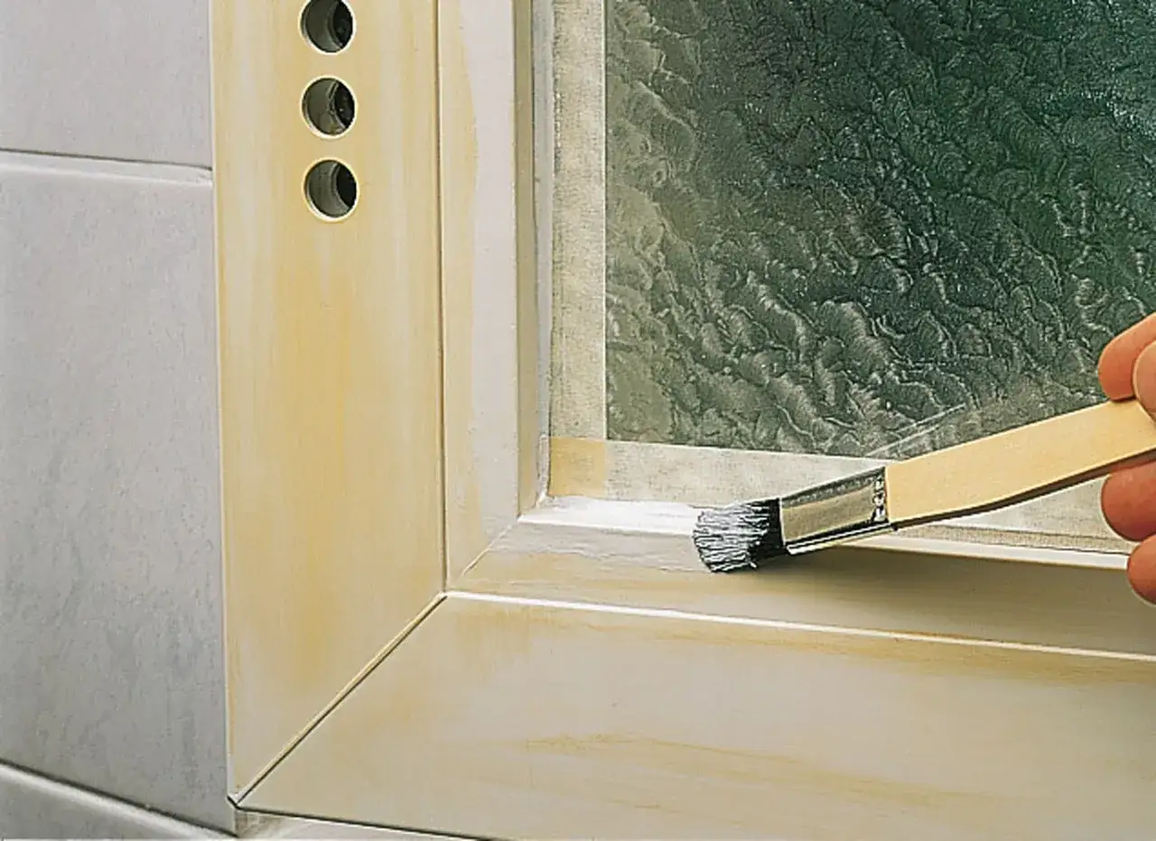 Hand streicht mit Pinsel über den Rahmen eines Kunststofffensters, um es zu renovieren.