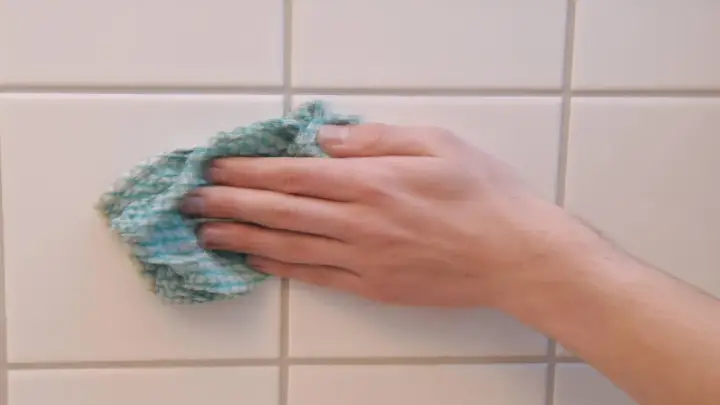 Hand wischt mit einem Tuch über weiße Wandfliesen, um sie zu reinigen.